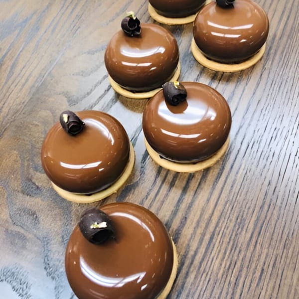 Six shiny, dome-shaped chocolate pastries with dark chocolate curls and gold flakes on a wooden surface.