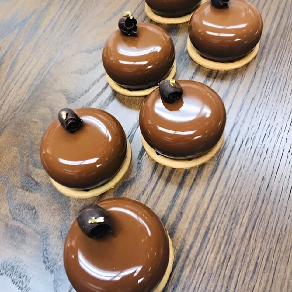 Six shiny, dome-shaped chocolate pastries with dark chocolate curls and gold flakes on a wooden surface.