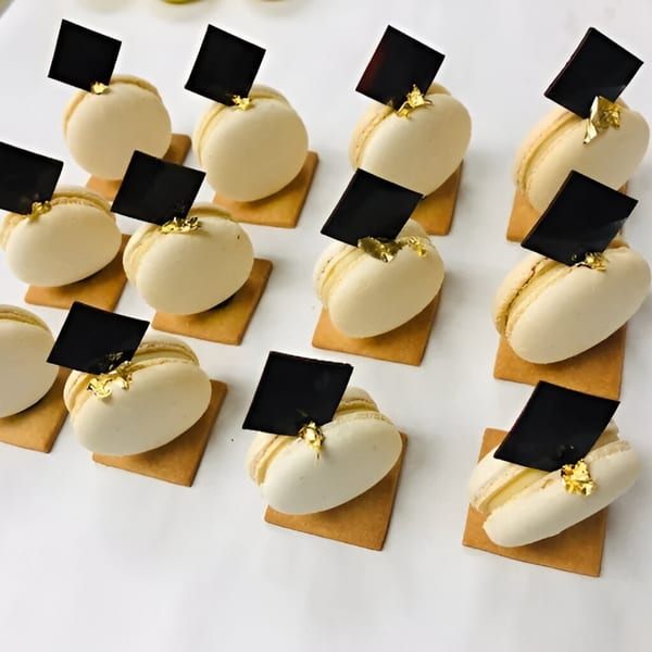 Rows of white macarons with gold flakes and black chocolate squares on small brown bases.