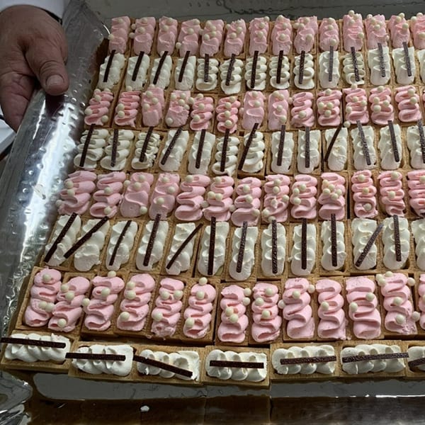 A hand holds a tray filled with rows of pink and white piped pastries, garnished with chocolate sticks and white pearls.