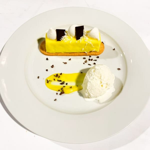Yellow glazed cake with white cream, chocolate squares, and a scoop of ice cream on a white plate.