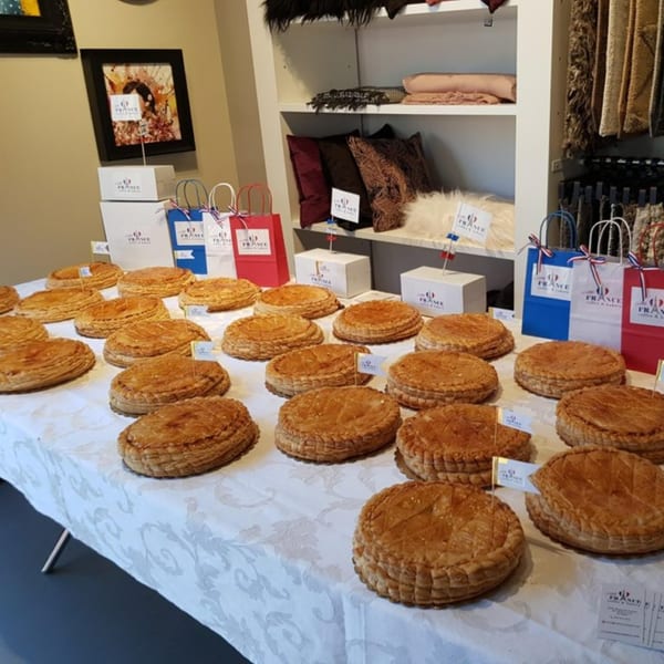 Many golden-brown Galette des Rois pastries with Little France flags on a white tablecloth.