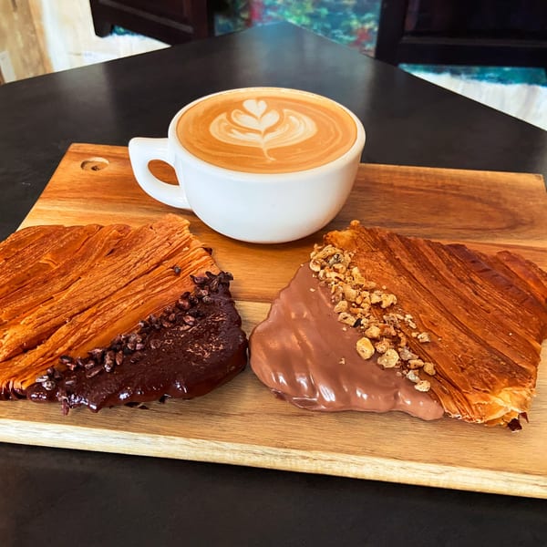 Two chocolate-dipped croissants and a latte with heart-shaped latte art on a wooden board.
