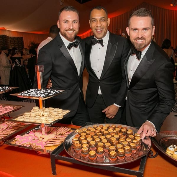 Three men in tuxedos smiling next to a dessert buffet with cookies and cupcakes.