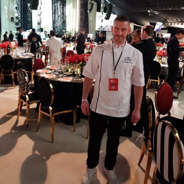 A chef in a white uniform stands in a large event space with set tables and other staff.