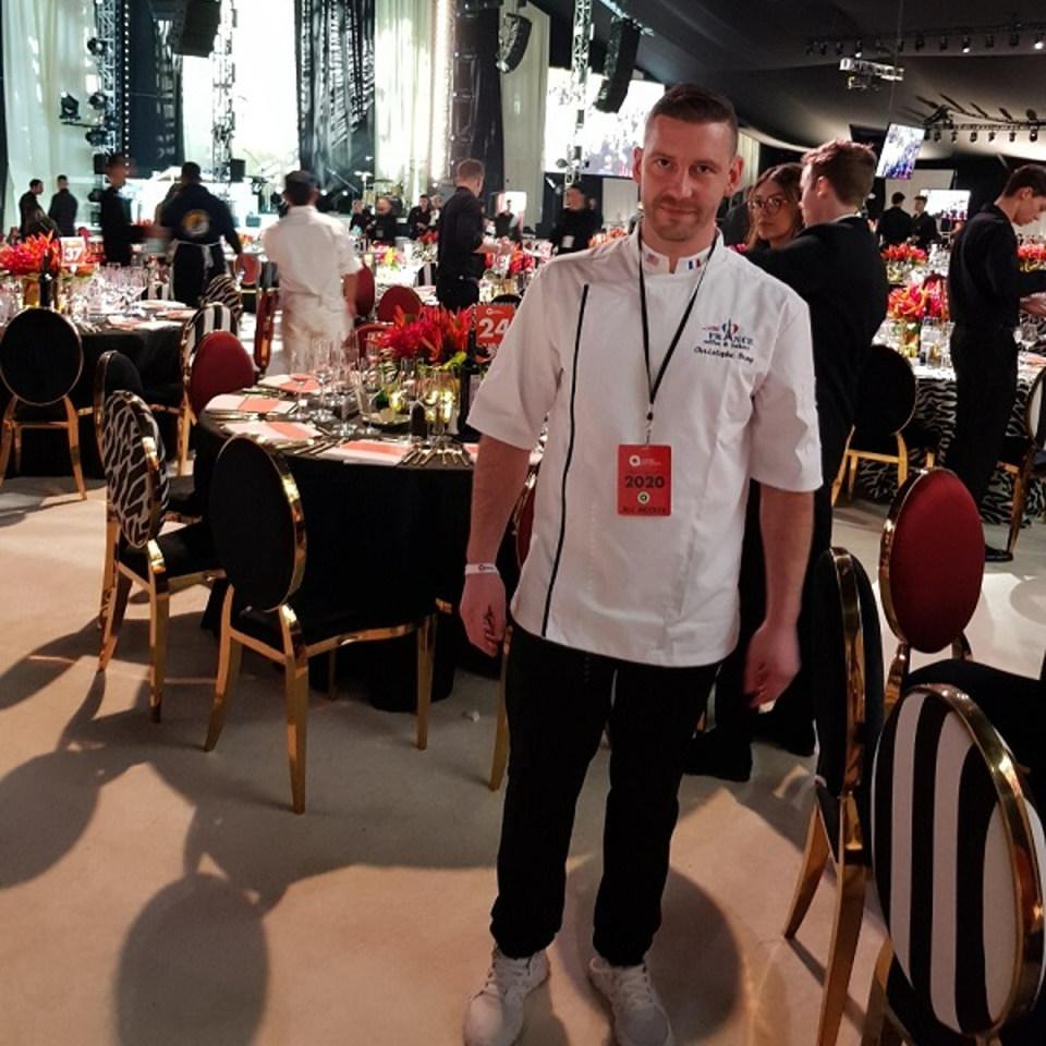 A chef in a white uniform stands in a large event space with set tables and other staff.