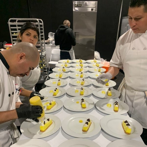 Chefs plating numerous yellow pastries with chocolate accents and sauce drops on white plates.