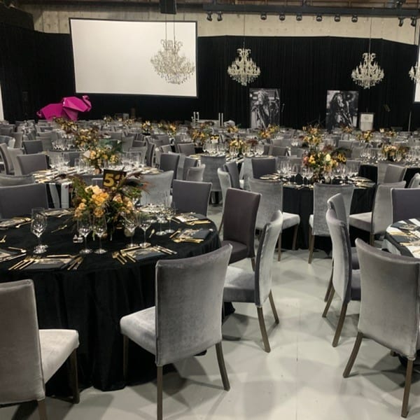 A large event space with numerous round tables set with black tablecloths, gold accents, and floral centerpieces.