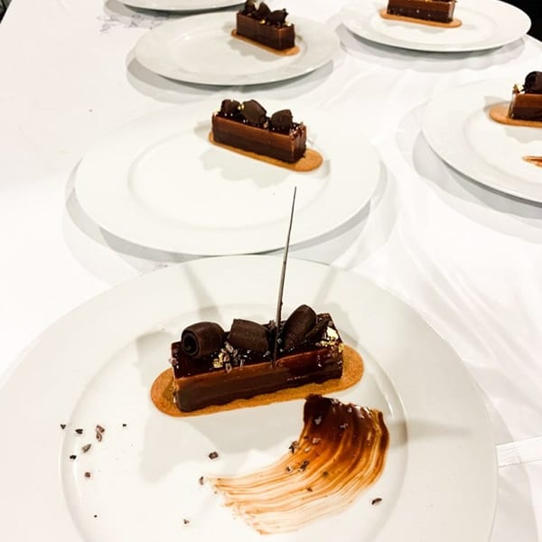 Multiple rectangular chocolate pastries with decorative toppings and sauce on white plates.