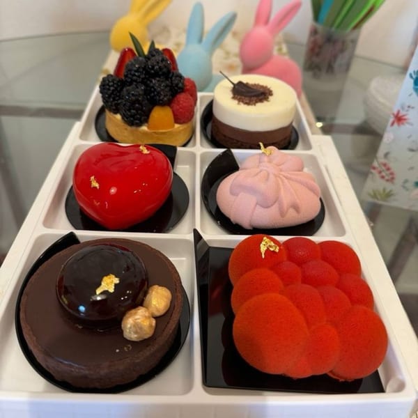 Six colorful, intricately decorated pastries, including fruit tarts and mousse cakes, in a white tray.