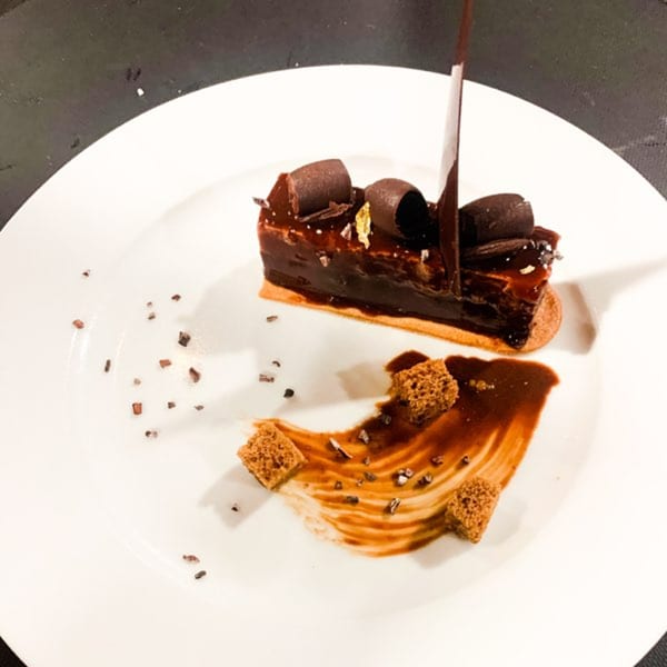 A chocolate eclair with chocolate curls and gold flakes, drizzled with chocolate sauce on a white plate.