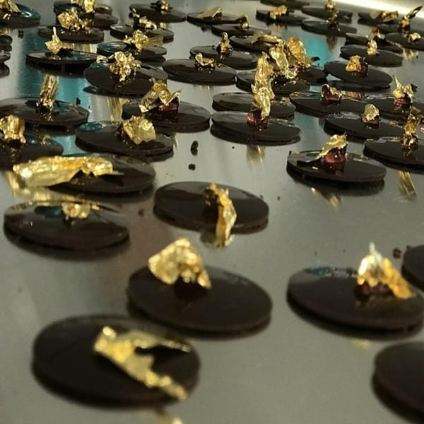 Rows of dark chocolate ovals topped with shimmering gold leaf on a reflective surface.