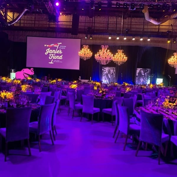 A large event space with purple lighting, round tables, chairs, chandeliers, and a projection screen.
