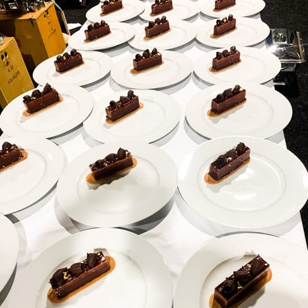 Rows of individual chocolate eclairs with chocolate shavings and gold flakes on white plates.