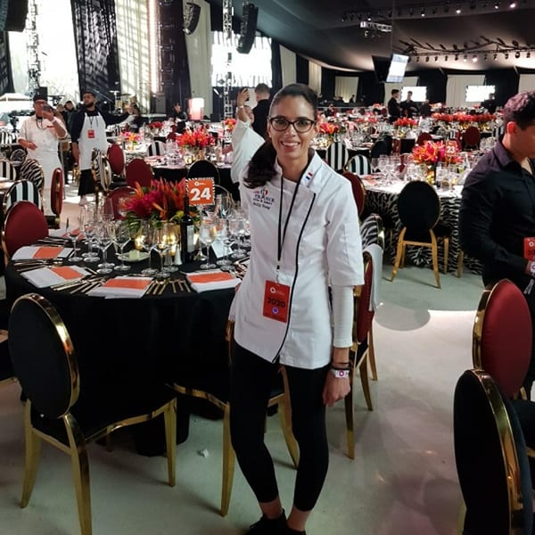 A chef in a white jacket smiles, standing in a large event space with decorated tables.