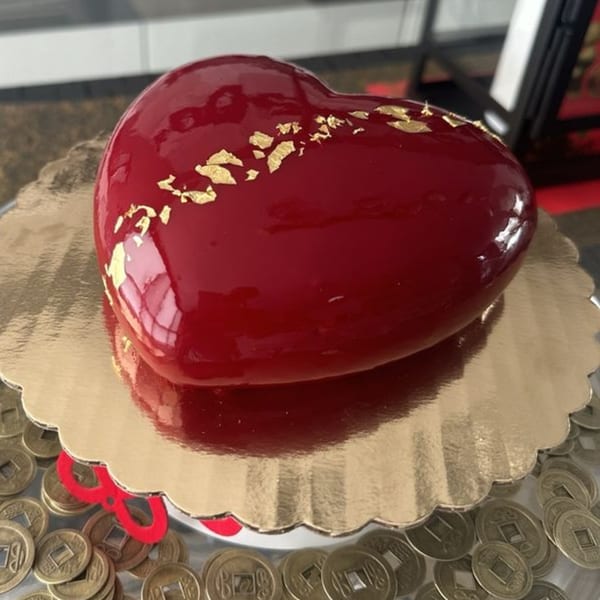 A heart-shaped red glazed cake with gold flakes sits on a gold scalloped tray.